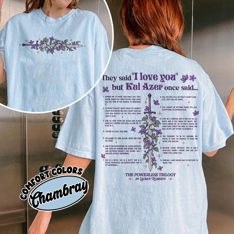 May include: Light blue t-shirt with a floral and sword design. The back of the shirt features text from "The Powerless Trilogy" by Lauren Roberts. The shirt is made of Comfort Colors Chambray fabric.