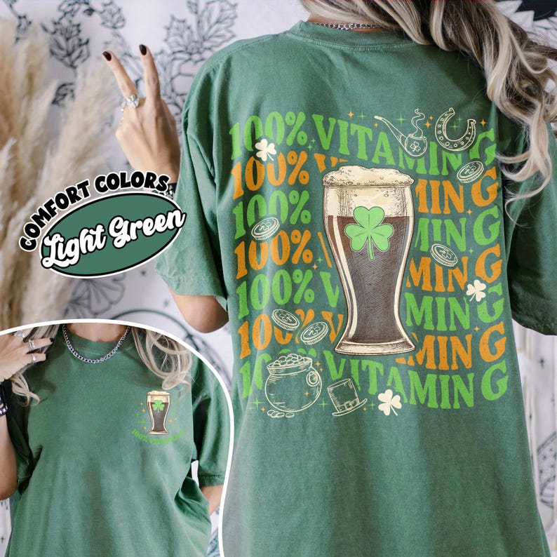 May include: Light green t-shirt with a St. Patrick's Day theme. The back features a large graphic of a beer glass with a shamrock, text that reads "100% Vitamin G", and other festive icons. The front has a smaller version of the beer glass graphic.