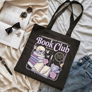 May include: Black tote bag with the text "UNAPOLOGETICALLY ANTI-SOCIAL Book Club" and a graphic of a skull, books, butterflies, and a teacup. The bag has long black handles and is lying on a cream-colored surface.