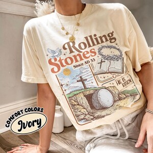 May include: Ivory-colored t-shirt with a graphic design featuring the words "Rolling Stones Since AD 33" and religious imagery. The design includes a cross, open book, and the phrase "He is Risen". The shirt is labeled "Comfort Colors Ivory".