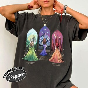 May include: A dark gray t-shirt with a graphic print of three women in stained glass windows. The text on the shirt reads "Let's go rattle the stars", "To the stars who listen and the dreams that are answered", and "Through love all is possible".