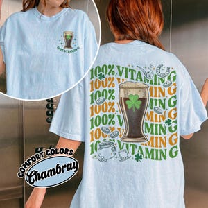 May include: Pale blue t-shirt with a St. Patrick's Day design. The front displays a beer glass graphic with a shamrock and the text "100% Vitamin G". The back has a larger design with the same theme, including a pot of gold and a horseshoe.