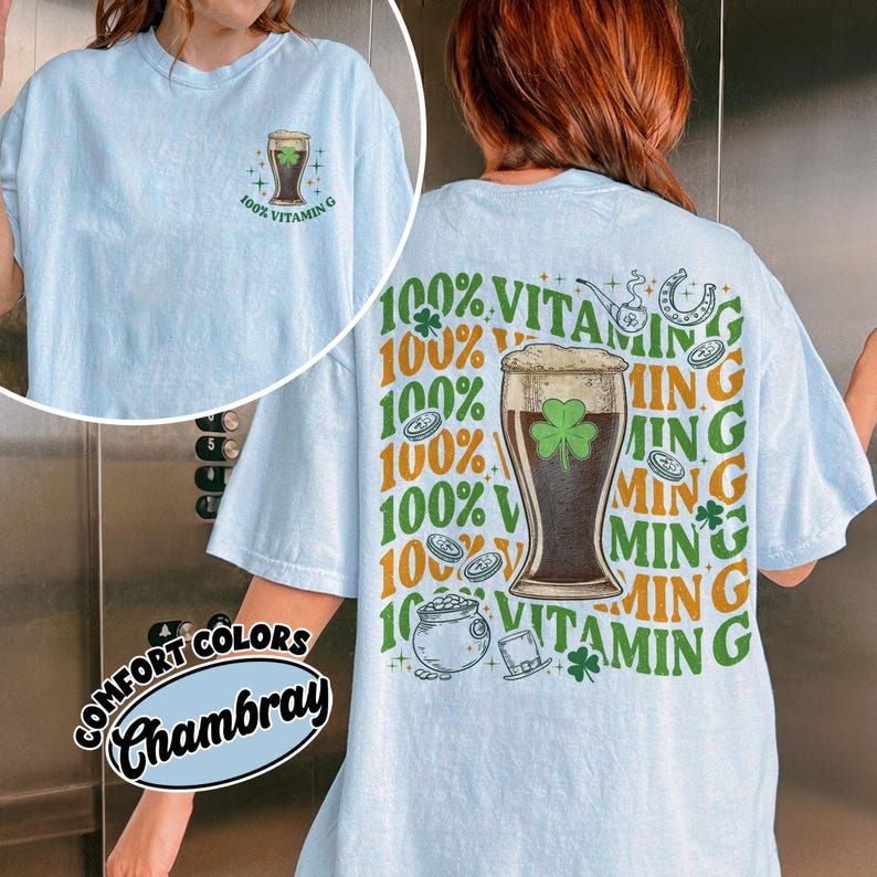May include: Pale blue t-shirt with a St. Patrick's Day design. The front displays a beer glass graphic with a shamrock and the text "100% Vitamin G". The back has a larger design with the same theme, including a pot of gold and a horseshoe.