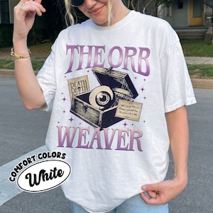 May include: White t-shirt with a graphic of a vintage wooden box with an eye inside. The text "THE ORB WEAVER" is printed in purple on the shirt. The text "DEATH DEATH" is printed on a small tag attached to the box.