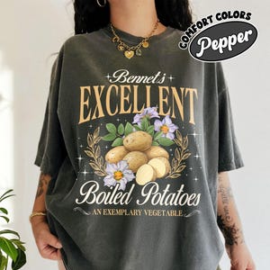 May include: A dark gray t-shirt with a graphic print of potatoes and flowers. The text on the shirt reads "Bennet's Excellent Boiled Potatoes An Exemplary Vegetable".