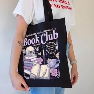 May include: Black tote bag with a graphic design featuring a skull, books, butterflies, and a teacup. Text on the bag reads "Unapologetically Anti-Social Book Club" and "Making Friends With Characters Not People."