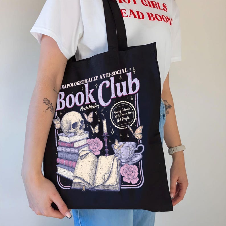 May include: Black tote bag with a graphic design featuring a skull, books, butterflies, and a teacup. Text on the bag reads "Unapologetically Anti-Social Book Club" and "Making Friends With Characters Not People."