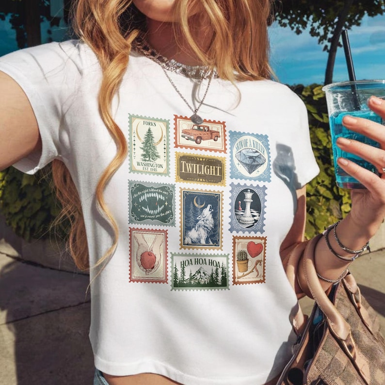 May include: White t-shirt featuring a collage of postage stamp designs. The stamps depict various images, including a red truck, a wolf howling at the moon, and the word "TWILIGHT". Other stamps show a lighthouse, a heart, and a tree.