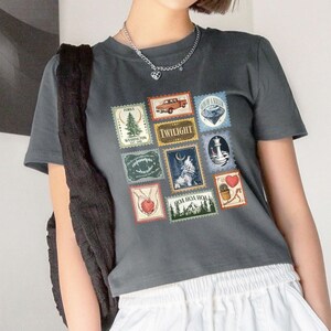 May include: Dark gray t-shirt featuring a collage of postage stamp-style graphics. The stamps include images of a truck, a wolf, a lighthouse, and the word "Twilight". The shirt has a crew neck and short sleeves.