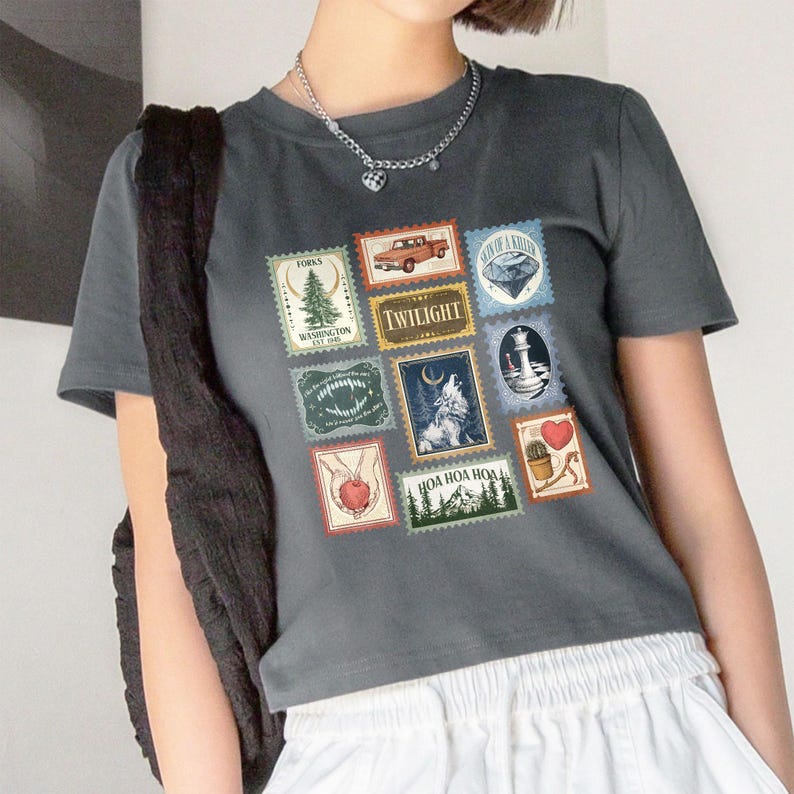 May include: Dark gray t-shirt featuring a collage of postage stamp-style graphics. The stamps include images of a truck, a wolf, a lighthouse, and the word "Twilight". The shirt has a crew neck and short sleeves.