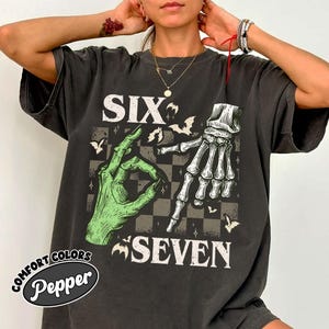 May include: Dark gray t-shirt with a graphic design featuring a green hand making an 'okay' sign and a white skeleton hand pointing down. The words "SIX" and "SEVEN" are above and below the hands, with bats and stars. The shirt has the words "COMFORT COLORS Pepper" on it.