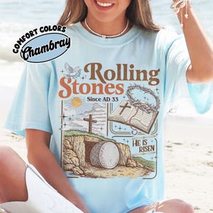 May include: Light blue t-shirt with a graphic featuring the words "Rolling Stones Since AD 33" and religious imagery. The design includes a cross, open book, and a tomb with a large stone. The shirt has the words "Comfort Colors Chambray" in a circle.