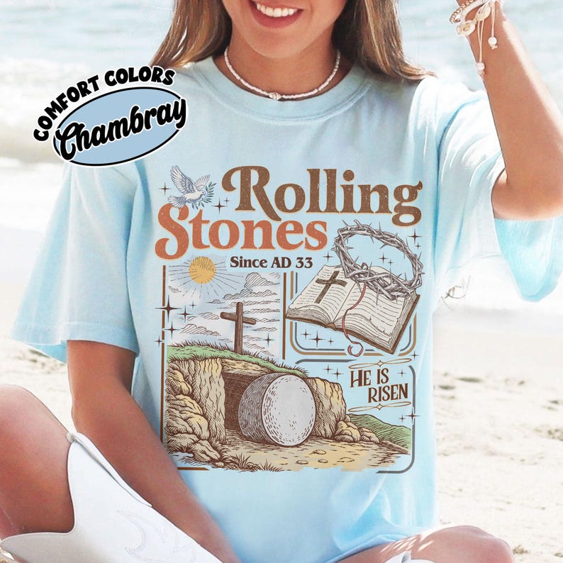 May include: Light blue t-shirt with a graphic featuring the words "Rolling Stones Since AD 33" and religious imagery. The design includes a cross, open book, and a tomb with a large stone. The shirt has the words "Comfort Colors Chambray" in a circle.