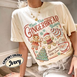 May include: Ivory-colored t-shirt with a festive "North Pole Gingerbread Bakery" graphic. The design features gingerbread people, a Christmas tree, and a Santa mug. The text "Handmade With Love" is also included.
