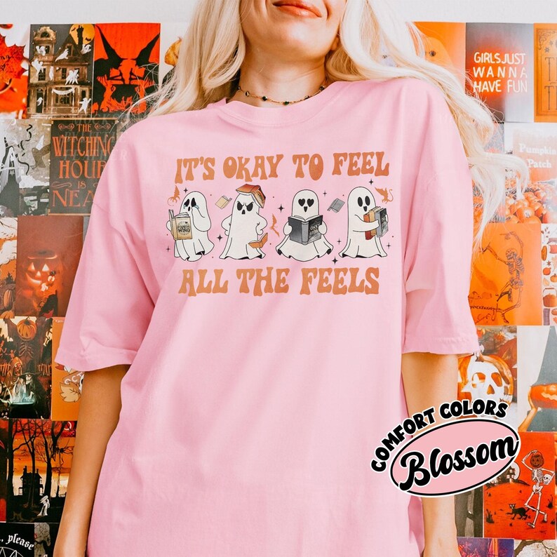May include: Pink t-shirt with a graphic of four ghosts reading books. The text "It's okay to feel all the feels" is printed on the shirt.