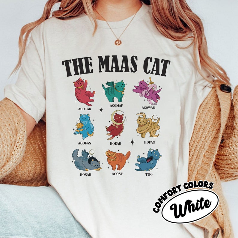 Puede incluir: Camiseta blanca con un gr&aacute;fico de nueve gatos en diferentes poses y colores. Cada gato tiene un nombre escrito debajo. El texto "THE MAAS CAT" est&aacute; en la parte superior del gr&aacute;fico. El texto "COMFORT COLORS White" est&aacute; en la parte inferior del gr&aacute;fico.