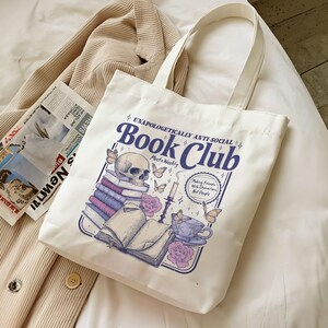 May include: Off-white canvas tote bag with the text "Book Club" in a vintage font. The graphic includes a skull, books, butterflies, a teacup, and the phrase "Making Friends with Characters, Not People."