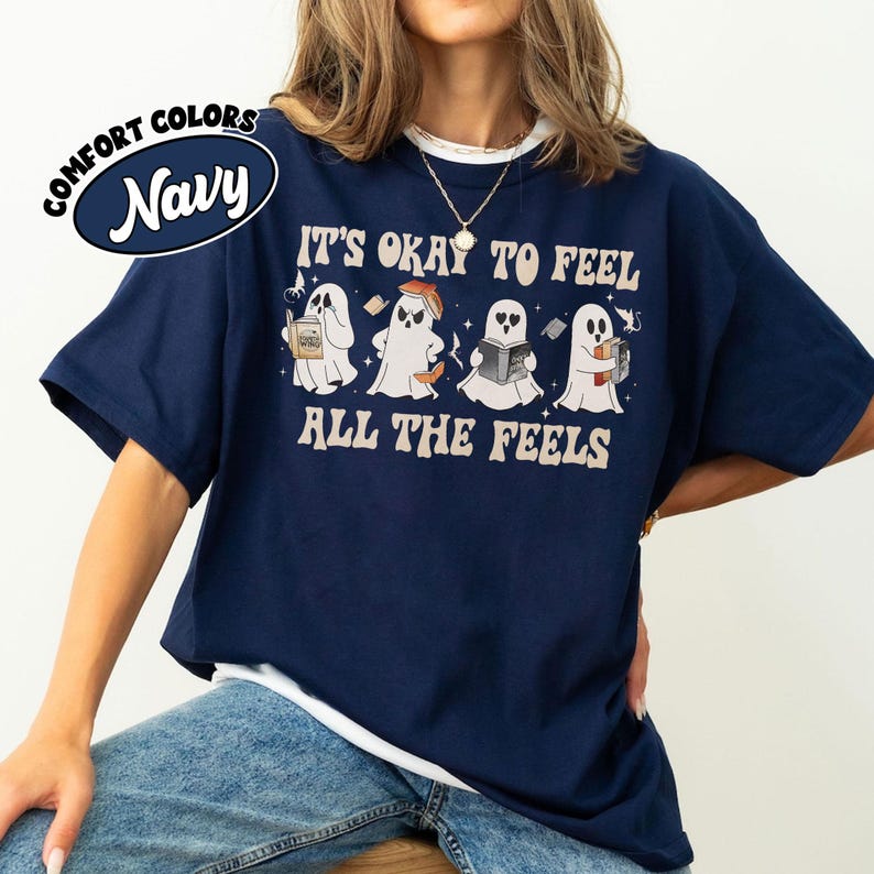 May include: Navy blue t-shirt with a white underlay. The shirt features a graphic of four ghosts reading books with the text "It's okay to feel all the feels".