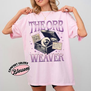 May include: Pink t-shirt with a graphic of an eye inside an open treasure chest with the text "The Orb Weaver" and "Death Death" on a tag. The text "Comfort Colors Blossom" is in a pink circle on the bottom left corner of the shirt.
