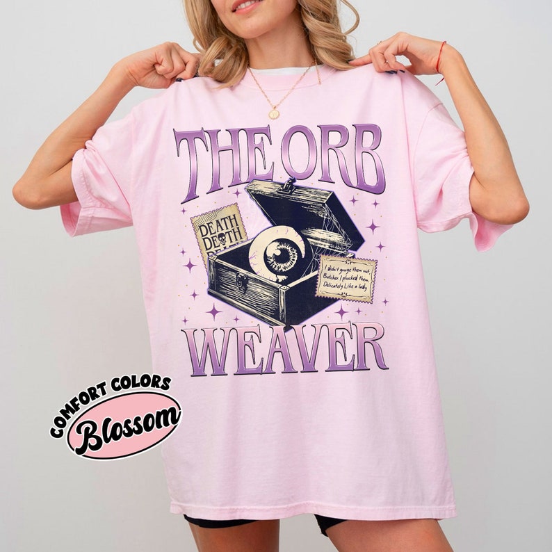 May include: Pink t-shirt with a graphic of an eye inside an open treasure chest with the text "The Orb Weaver" and "Death Death" on a tag. The text "Comfort Colors Blossom" is in a pink circle on the bottom left corner of the shirt.