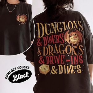 May include: Black t-shirt with a graphic featuring a dragon, dice, and the text "DUNGEONS & DINERS & DRAGONS & DRIVE-INS & DIVES." The shirt also has a small dragon and dice graphic on the front. The words "COMFORT COLORS" and "Black" are also visible.