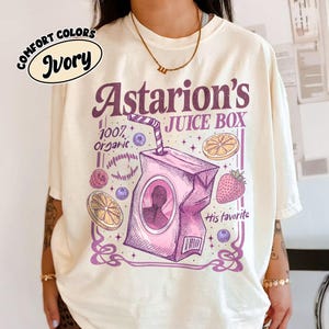 May include: Ivory-colored t-shirt with a graphic design. The design features the text "Astarion's JUICE BOX" in purple, along with illustrations of fruit and a juice box. The words "100% organic" are also included.