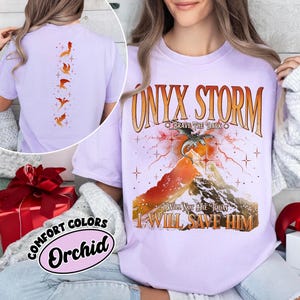 May include: A light purple t-shirt with the text "ONYX STORM" and a graphic of a dragon over a mountain. The shirt also features the phrases "BRAVE THE DARK", "I WILL NOT DIE TODAY", and "I WILL SAVE HIM". The back of the shirt has a design of orange dragons.