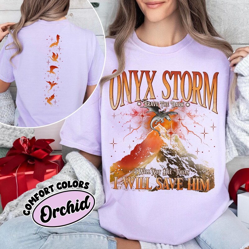 May include: A light purple t-shirt with the text "ONYX STORM" and a graphic of a dragon over a mountain. The shirt also features the phrases "BRAVE THE DARK", "I WILL NOT DIE TODAY", and "I WILL SAVE HIM". The back of the shirt has a design of orange dragons.