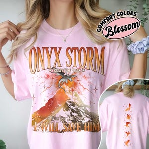 May include: A light pink t-shirt with the words "ONYX STORM" in an orange, distressed font. Below the text is a graphic of a dragon over a mountain. The text "BRAVE THE DARK" and "I WILL NOT DIE TODAY I WILL SAVE HIM" are also printed on the shirt. The back of the shirt has a vertical design of orange dragons.