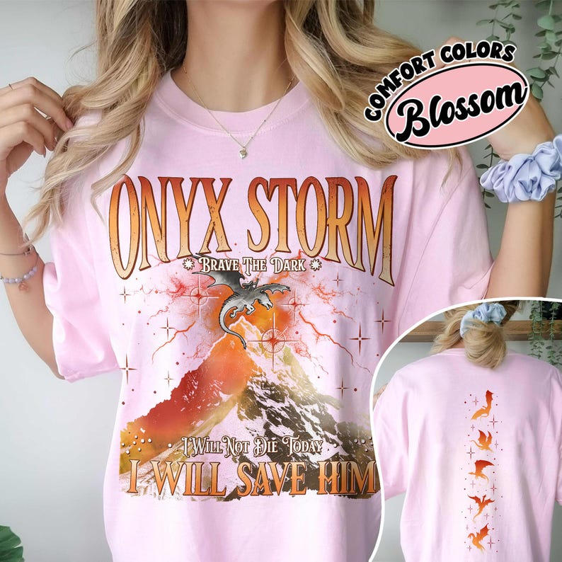 May include: A light pink t-shirt with the words "ONYX STORM" in an orange, distressed font. Below the text is a graphic of a dragon over a mountain. The text "BRAVE THE DARK" and "I WILL NOT DIE TODAY I WILL SAVE HIM" are also printed on the shirt. The back of the shirt has a vertical design of orange dragons.