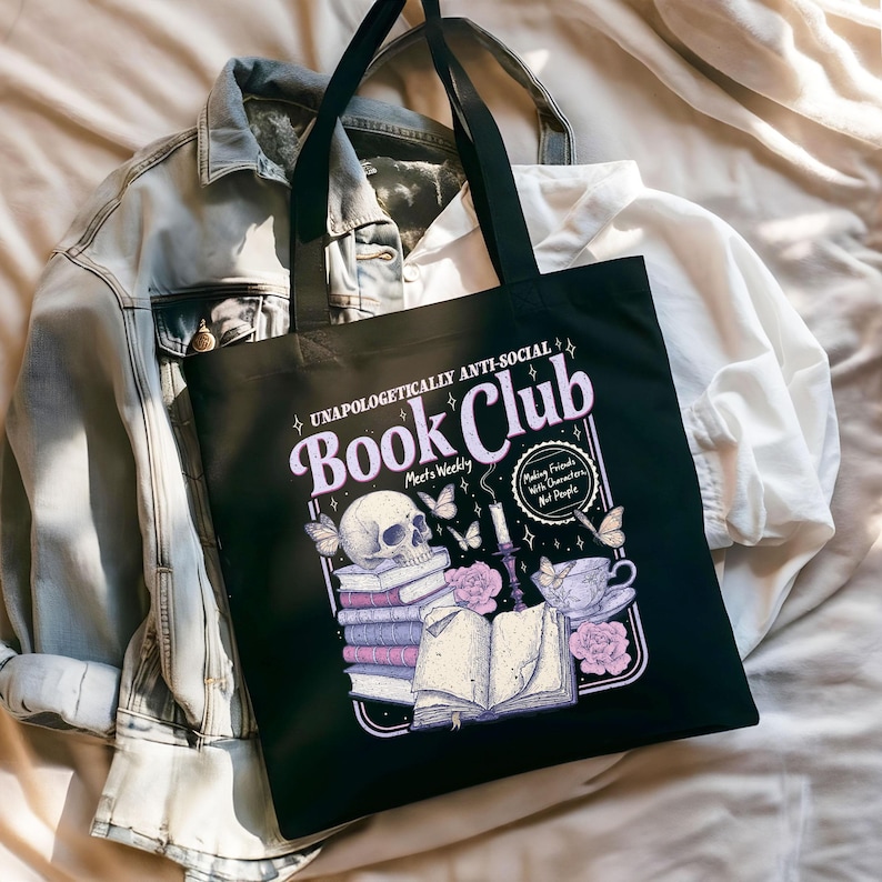 May include: Black tote bag with the text "Book Club" in a light purple font. The bag features a skull, books, butterflies, flowers, and a teacup graphic. The text "Unapologetically Anti-Social" and "Making Friends With Characters, Not People" are also included.