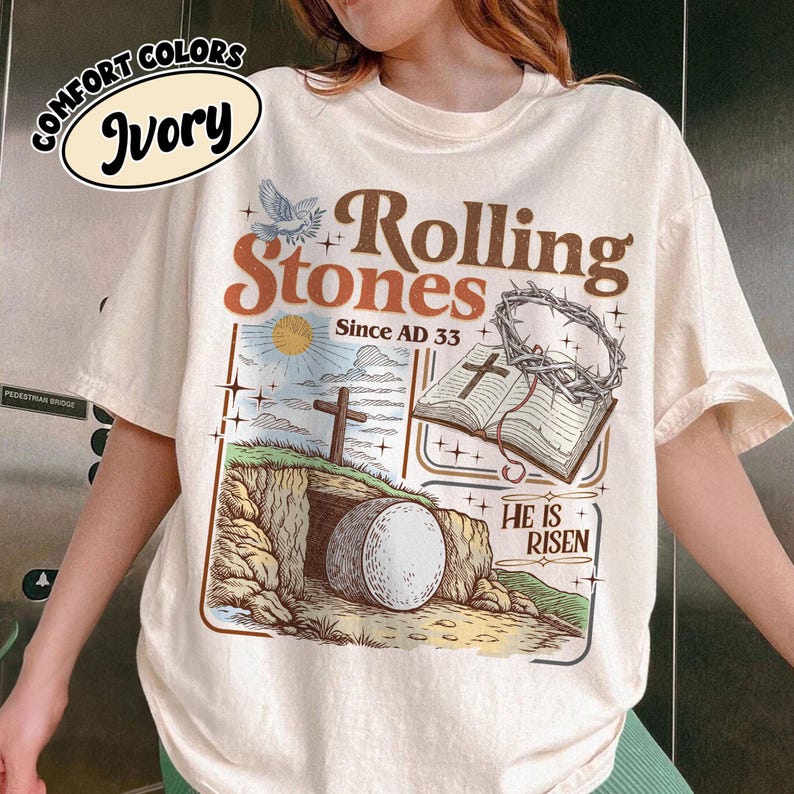 May include: Ivory-colored t-shirt with a graphic design featuring the words "Rolling Stones Since AD 33." The design includes a cross, an open book, and a tomb with a large stone. The shirt also has the words "He is Risen."