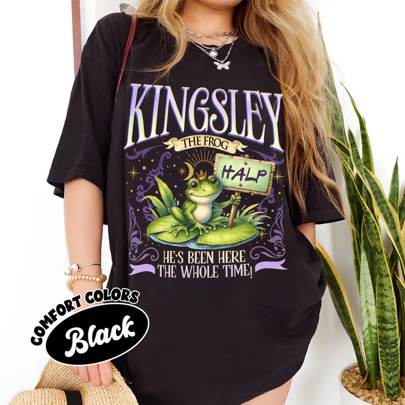 Puede incluir: Camiseta negra con un gr&aacute;fico de una rana llamada Kingsley en una hoja de nen&uacute;far, sosteniendo un letrero que dice "HALP". El dise&ntilde;o incluye el texto "KINGSLEY THE FROG" y "HE'S BEEN HERE THE WHOLE TIME!" La camiseta tambi&eacute;n tiene el texto "COMFORT COLORS Black".