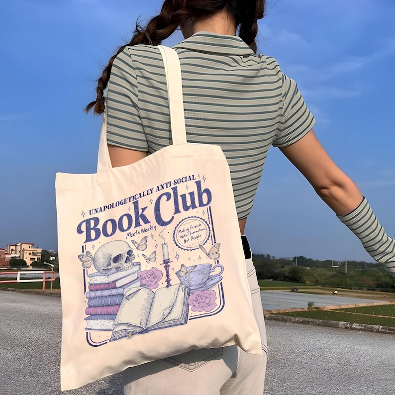 May include: Cream-colored tote bag with a "Book Club" graphic. The design includes a skull, books, butterflies, a teacup, and the text "Unapologetically Anti-Social." The bag has a cream-colored strap.