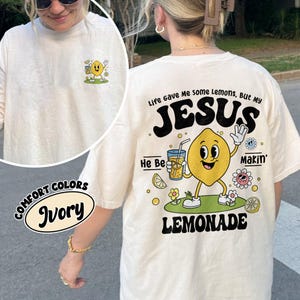 May include: Ivory-colored t-shirt with a cartoon lemon character holding a lemonade drink. The back of the shirt features the text "Life Gave Me Some Lemons, But My JESUS He Be Makin' Lemonade."