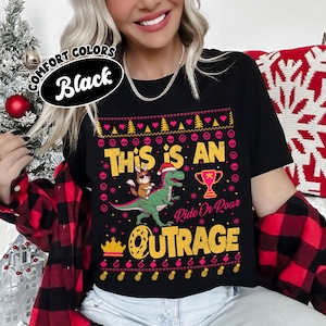 May include: Black t-shirt with a festive design. The shirt features the text "THIS IS AN OUTRAGE" with a dinosaur wearing a Santa hat and a bunny riding it. The design includes a trophy, hearts, and Christmas trees.