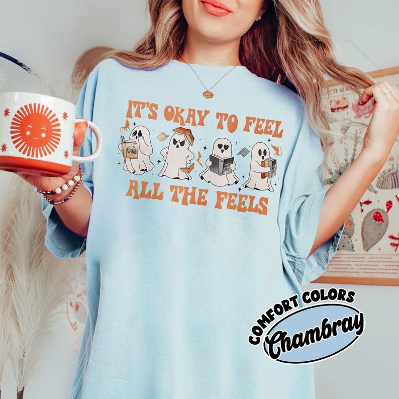 May include: Light blue t-shirt with a graphic of four ghosts reading books. The text "It's okay to feel all the feels" is printed on the shirt. The shirt is made by Comfort Colors Chambray.