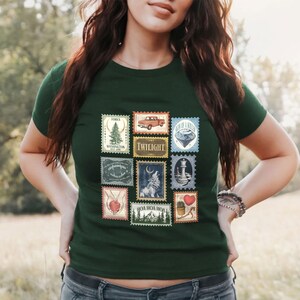 May include: Dark green t-shirt featuring a collection of postage stamp designs. The stamps depict various images, including a pine tree, a red truck, a wolf, and the word "TWILIGHT".