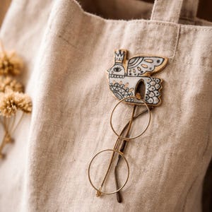 May include: A beige linen tote bag with a decorative bird pin and gold-rimmed glasses. The bird pin is white with black and blue details, and a small crown. The glasses have round lenses.