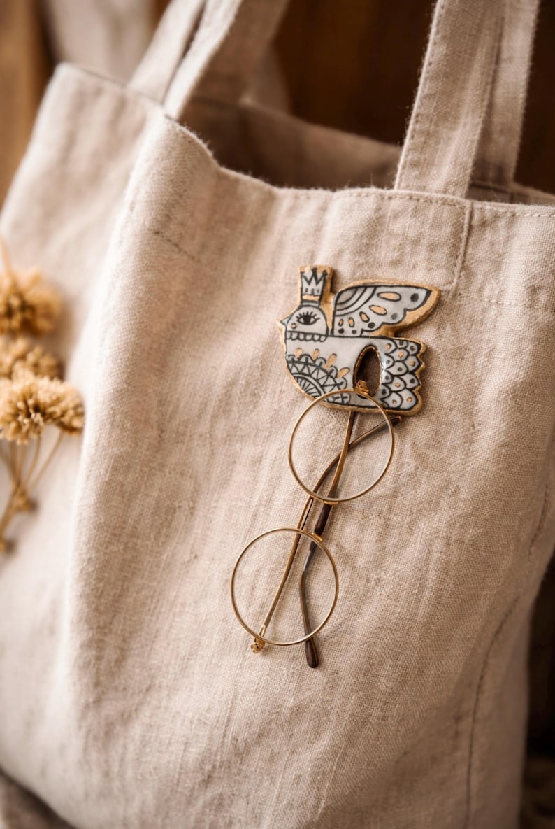 May include: A beige linen tote bag with a decorative bird pin and gold-rimmed glasses. The bird pin is white with black and blue details, and a small crown. The glasses have round lenses.