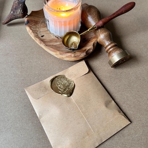 May include: A lit candle in a glass jar sits on a wooden tray with a small wooden bird. A brass wax melting spoon and a wooden handled wax stamp are next to the candle. A brown paper envelope is sealed with a gold wax seal.