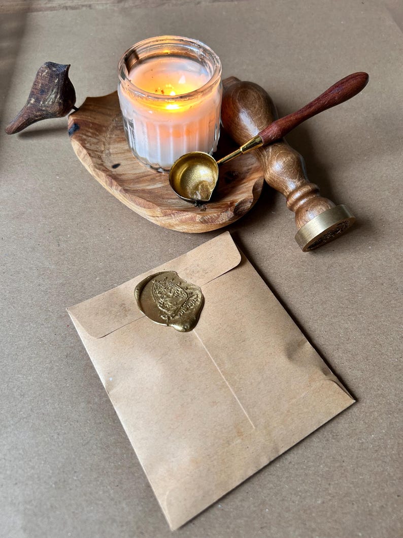 May include: A lit candle in a glass jar sits on a wooden tray with a small wooden bird. A brass wax melting spoon and a wooden handled wax stamp are next to the candle. A brown paper envelope is sealed with a gold wax seal.