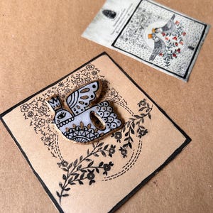 May include: A handmade bird pin with a crown, wings, and decorative details, set on a brown card with black floral accents. A second card with bird illustrations is visible in the background.