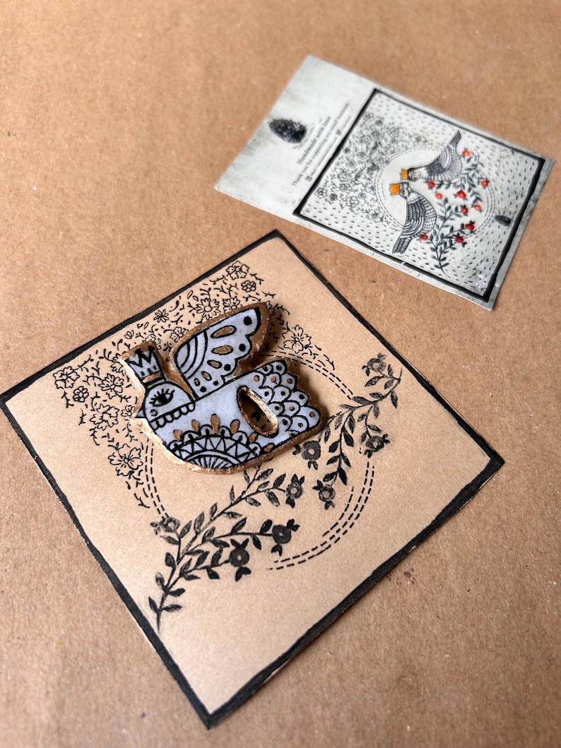 May include: A handmade bird pin with a crown, wings, and decorative details, set on a brown card with black floral accents. A second card with bird illustrations is visible in the background.