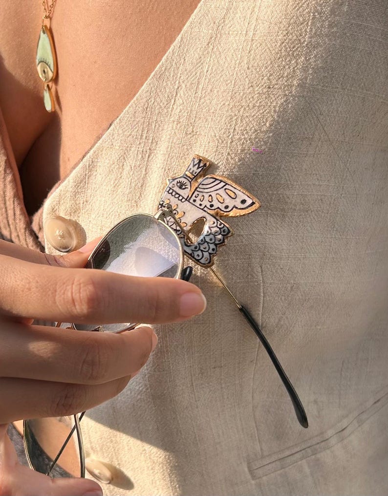 May include: A decorative butterfly pin with black and white details is attached to a beige vest. The pin is paired with a pair of sunglasses with a gold frame. A gold necklace with a teardrop pendant is also visible.