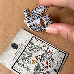 May include: A white and gold bird-shaped ceramic brooch with black detailing and a crown. The brooch is held by a hand. A card with a bird illustration and text is in the background.