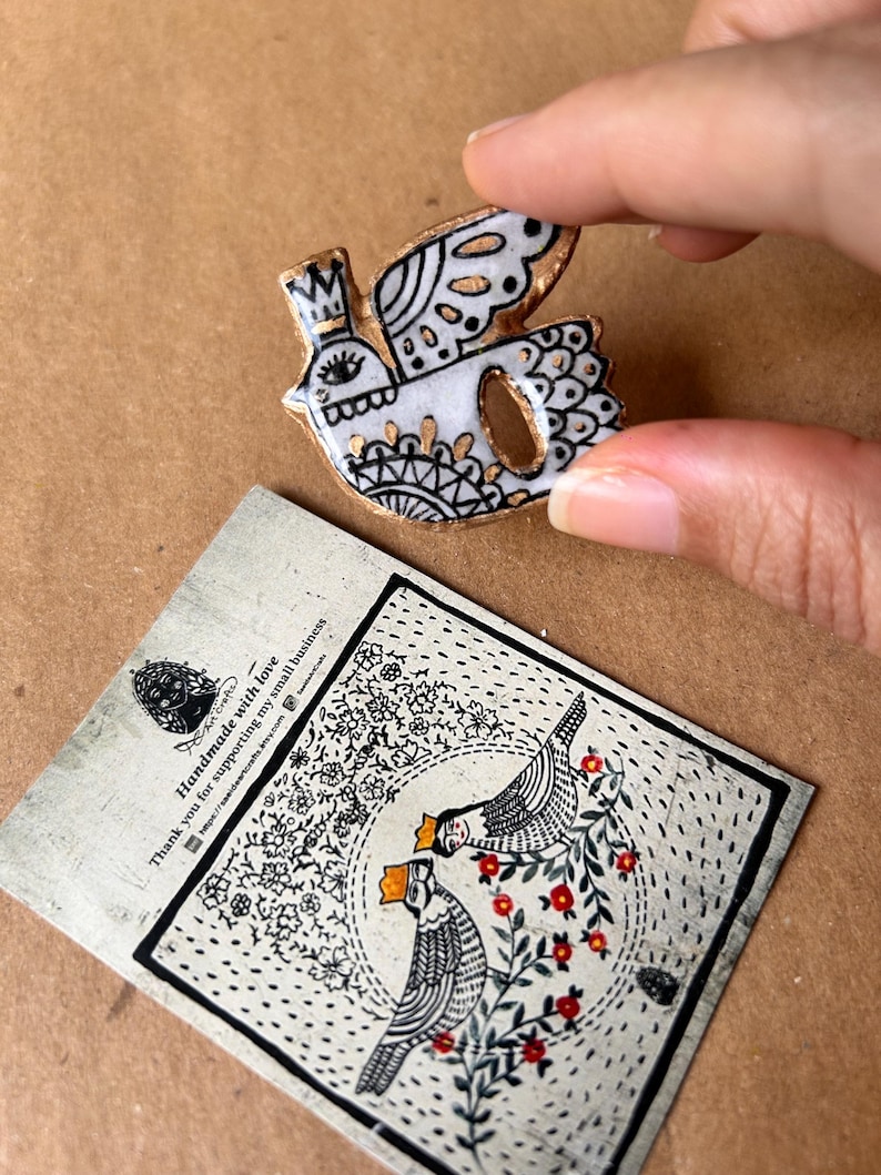 May include: A white and gold bird-shaped ceramic brooch with black detailing and a crown. The brooch is held by a hand. A card with a bird illustration and text is in the background.