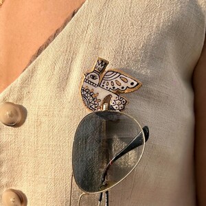 May include: A decorative bird-shaped pin with a crown, eye, and wing details. The pin is white with black and gold accents. Sunglasses are clipped to the pin. The pin is attached to a beige vest.