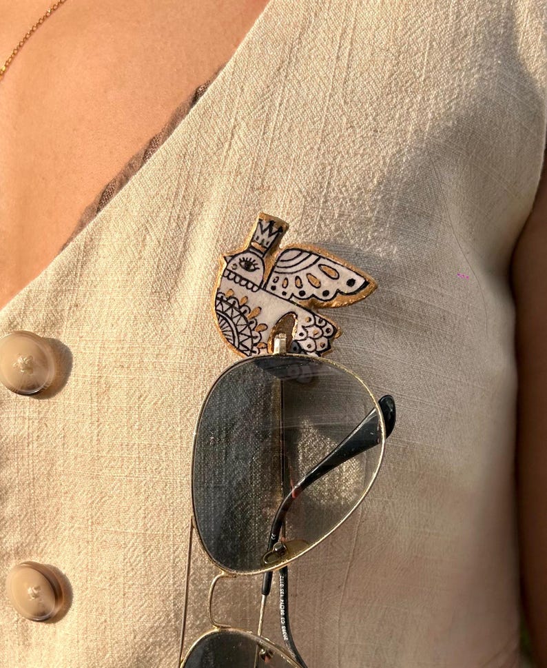 May include: A decorative bird-shaped pin with a crown, eye, and wing details. The pin is white with black and gold accents. Sunglasses are clipped to the pin. The pin is attached to a beige vest.