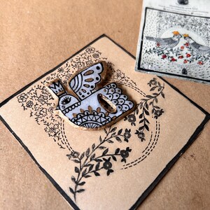 May include: A decorative bird-shaped button with a crown, featuring white, gold, and black details, rests on a diamond-shaped card with floral and leaf patterns. A small card with two birds and floral details is also present.